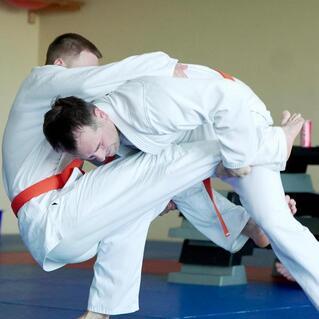 Zwei Judokas in wei&szlig;en Anz&uuml;gen f&uuml;hren eine W&uuml;rgetechnik auf einer Matte aus, w&auml;hrend Zuschauer im Hintergrund sitzen.