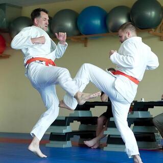 Zwei Judokas in wei&szlig;en Anz&uuml;gen &uuml;ben einen Kick auf einer blauen Matte, w&auml;hrend Zuschauer im Hintergrund sitzen.