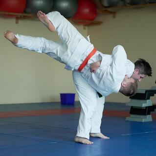 Zwei Judoka in wei&szlig;en Anz&uuml;gen f&uuml;hren einen Wurf im Training auf matten Boden aus. Der eine hat einen orangefarbenen G&uuml;rtel.