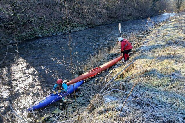 Zwei Personen entladen Kanus am Ufer eines schmalen, glitzernden Gew&auml;ssers in einer bewaldeten Umgebung.