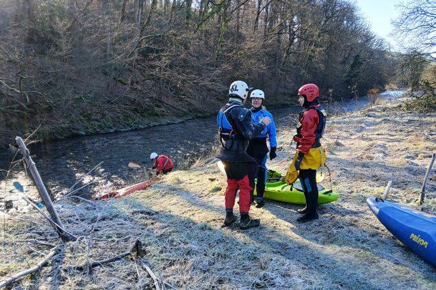 Vier Paddler in Neoprenanz&uuml;gen stehen am Ufer eines gefrorenen Flusses, w&auml;hrend ein weiteres Boot im Wasser liegt.