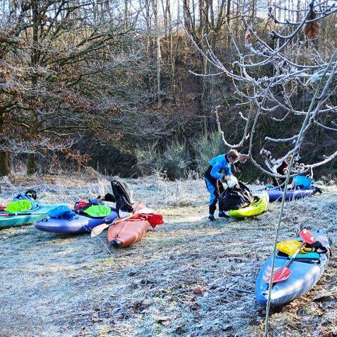 Zwei Personen packen Kajaks auf frostigem Ufer, umgeben von B&auml;umen und einer unber&uuml;hrten Winterlandschaft.