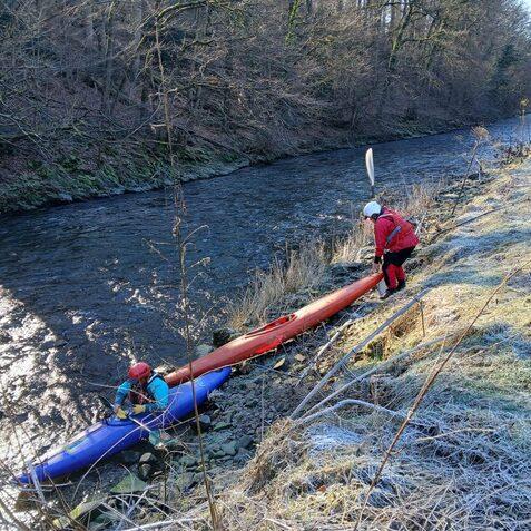 Zwei Personen entladen Kanus am Ufer eines schmalen, glitzernden Gew&auml;ssers in einer bewaldeten Umgebung.