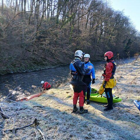 Vier Paddler in Neoprenanz&uuml;gen stehen am Ufer eines gefrorenen Flusses, w&auml;hrend ein weiteres Boot im Wasser liegt.