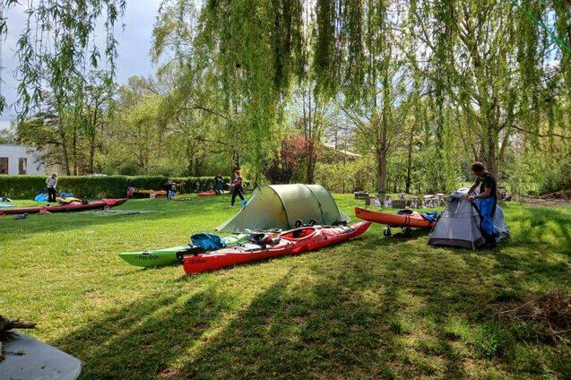Zelt und Kajaks auf einer gr&uuml;nen Wiese, umgeben von B&auml;umen; Personen bereiten Ausr&uuml;stung f&uuml;r eine Outdoor-Aktivit&auml;t vor.