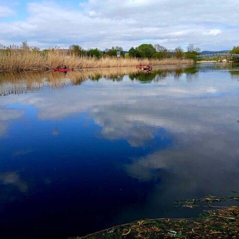 Ruhiger Gew&auml;sser mit Spiegelung des Himmels und Uferbewuchs, zwei Kanufahrer paddeln in der Ferne.