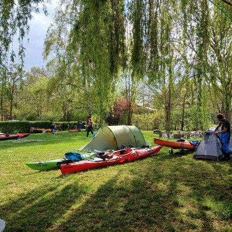Zelt und Kajaks auf einer gr&uuml;nen Wiese, umgeben von B&auml;umen; Personen bereiten Ausr&uuml;stung f&uuml;r eine Outdoor-Aktivit&auml;t vor.