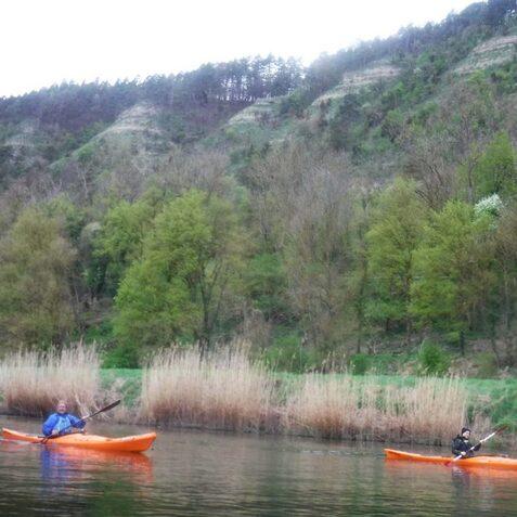 Zwei Kajakfahrer in orangen Booten paddeln auf einem ruhigen Gew&auml;sser, umgeben von gr&uuml;nen B&auml;umen und h&uuml;geliger Landschaft.