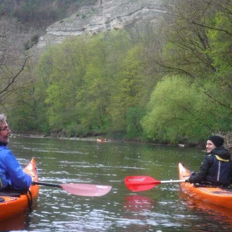 Zwei Personen in orangefarbenen Kajaks auf einem ruhigen Gew&auml;sser, umgeben von gr&uuml;nen B&auml;umen und Felsen.