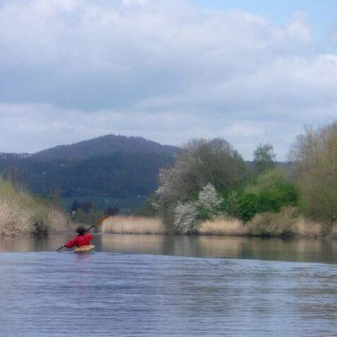 Kanufahrer in rotem Boot paddelt auf ruhigem Wasser, umgeben von B&auml;umen und sanften H&uuml;geln unter einem bew&ouml;lkten Himmel.