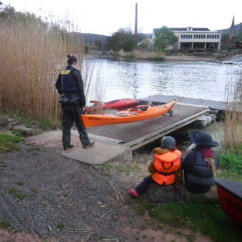 Person mit Kajak auf einem Steg, w&auml;hrend zwei Kinder in Schwimmwesten am Ufer sitzen und das Wasser beobachten.