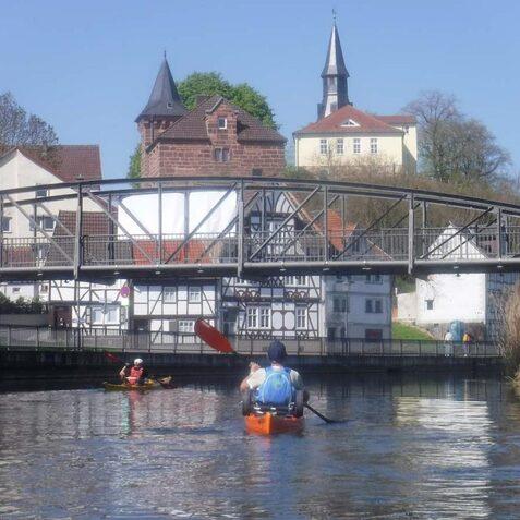 Zwei Kajakfahrer paddeln auf einem Fluss unter einer Metallbr&uuml;cke mit H&auml;usern im Hintergrund und einer Kirche.