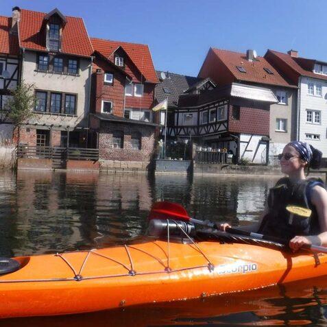 Frau im Kanu auf einem ruhigen Gew&auml;sser mit historischen H&auml;usern im Hintergrund und strahlend blauem Himmel.