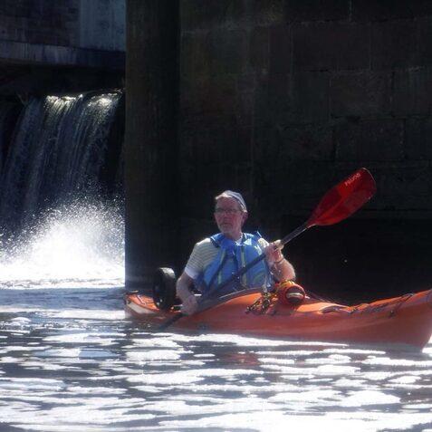 Mann im Kajak paddelt auf einem Gew&auml;sser, neben einem Wasserfall und einer senkrechten Mauer.