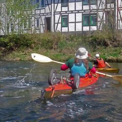 Zwei Kajakfahrer paddeln auf einem ruhigen Fluss, w&auml;hrend ein Fachwerkhaus im Hintergrund sichtbar ist.