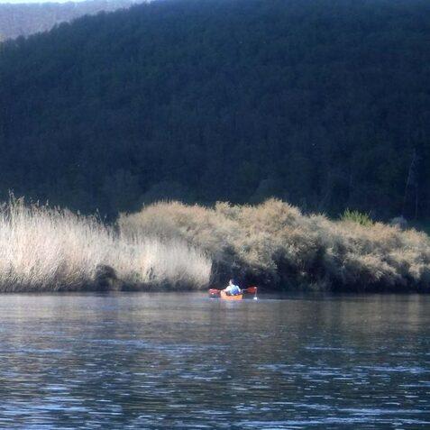 Kajakfahrer auf einem stillen Gew&auml;sser, umgeben von Schilf und bewaldeten H&uuml;geln im Hintergrund.