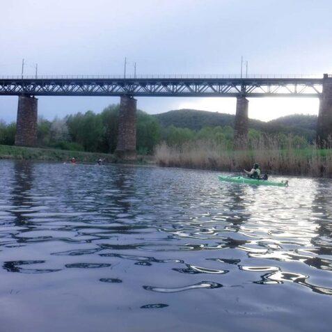 Kaum sichtbare Kajakfahrer auf einem ruhigen Gew&auml;sser, im Hintergrund eine typische Steinbr&uuml;cke und bewaldete H&uuml;gel.