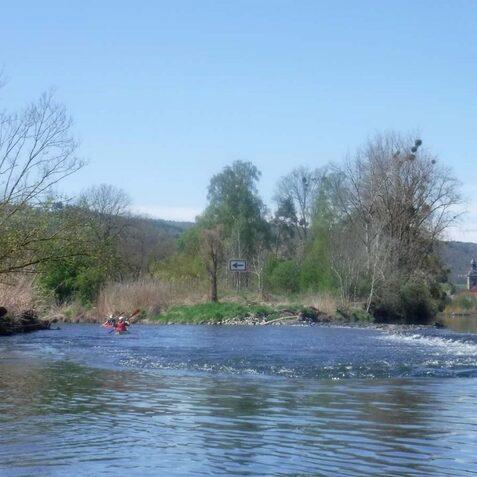 Zwei Kanufahrer paddeln auf einem ruhigen Fluss, umgeben von B&auml;umen und sanften H&uuml;geln im Hintergrund.