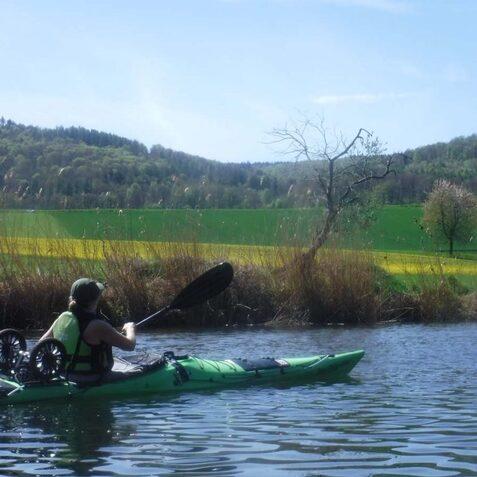 Paddelndes Kajak auf einem ruhigen Gew&auml;sser, mit einer Person in sportlicher Kleidung vor einer gr&uuml;nen, bewaldeten Landschaft.