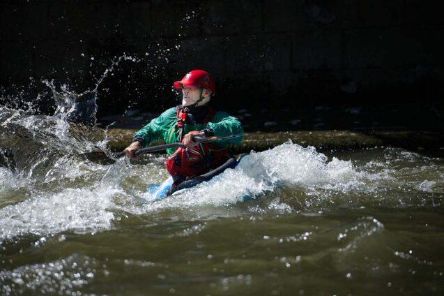 Kajakfahrer paddelt durch spritzende Wellen in einem Fluss, getragen von einer gr&uuml;nen Weste und rotem Helm.