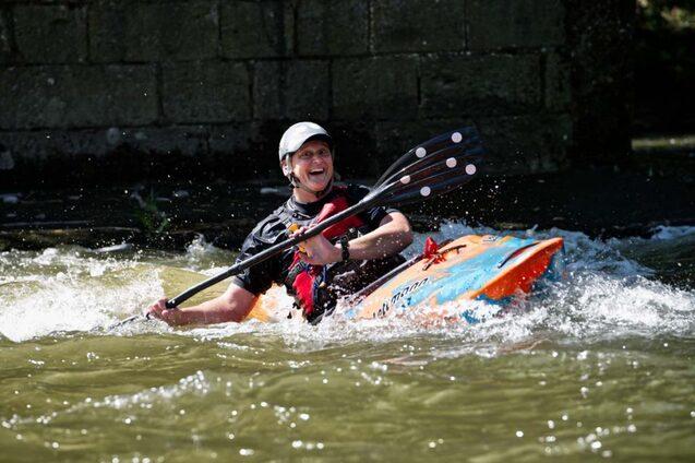 Person in farbenfrohem Kajak paddelt l&auml;chelnd durch sprudelndes Wasser neben einer Steinmauer.