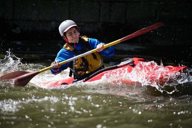 Junger Kanute paddelt in einem roten Kajak durch spritzende Wellen auf einem Fluss.