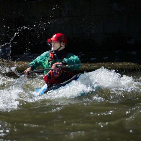 Kajakfahrer paddelt durch spritzende Wellen in einem Fluss, getragen von einer gr&uuml;nen Weste und rotem Helm.