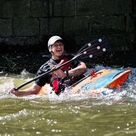Person in farbenfrohem Kajak paddelt l&auml;chelnd durch sprudelndes Wasser neben einer Steinmauer.