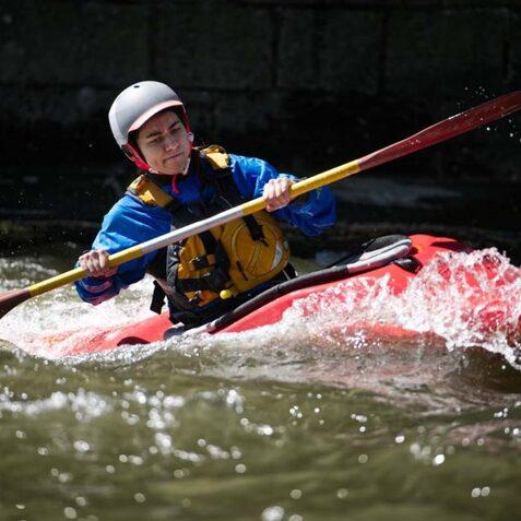 Junger Kanute paddelt in einem roten Kajak durch spritzende Wellen auf einem Fluss.
