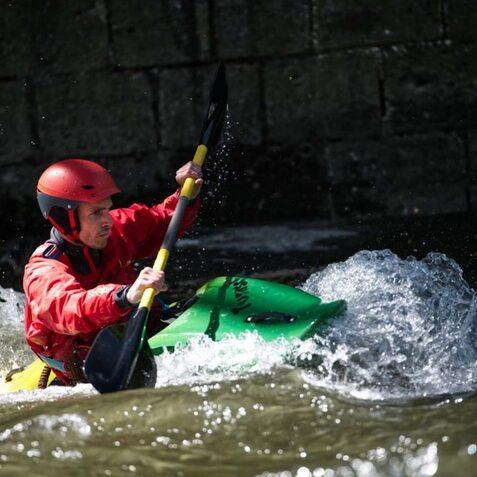 Ein Kajakfahrer in roter Kleidung paddelt mit einem gr&uuml;nen Kajak durch unruhige Gew&auml;sser.