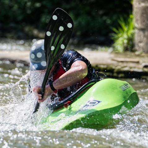 Kajakfahrer paddelt energisch in einem spritzendem Wasser auf einem ruhigen Fluss.