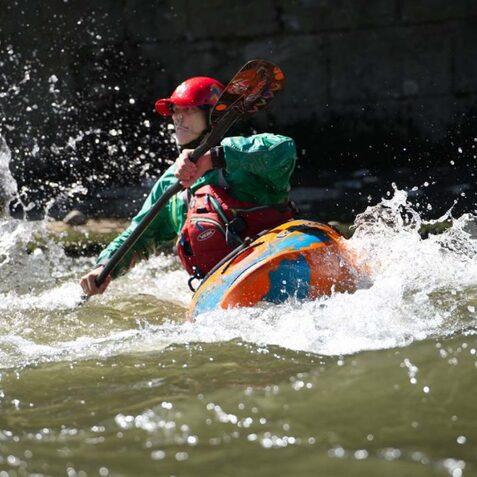 Kind paddelt in einem bunten Kajak durch spritzige Wellen eines Flusses an einem sonnigen Tag.