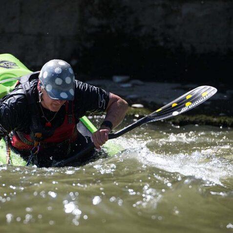 Paddler in einem gr&uuml;nen Kajak k&auml;mpft im Wasser, w&auml;hrend er mit einem Paddel rudert. Sonne reflektiert auf der Wasseroberfl&auml;che.