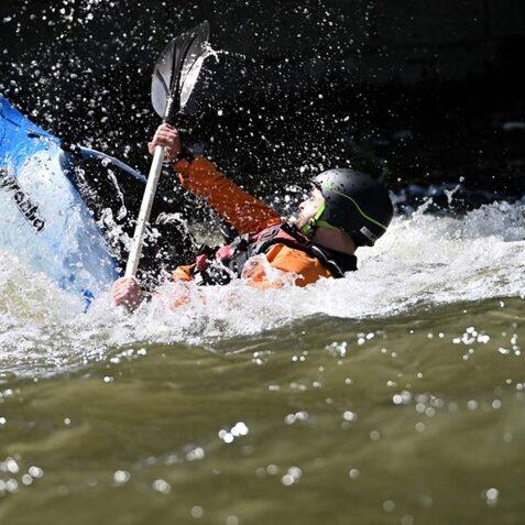 Kajakfahrer in einem blauen Kajak paddelt in unruhigem Wasser, w&auml;hrend spritzende Wellen um ihn herum aufsteigen.