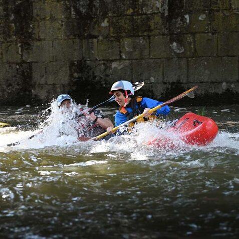 Zwei Personen im Kanu k&auml;mpfen gegen Wellen in einem Fluss, w&auml;hrend Wasser &uuml;ber sie spritzt.