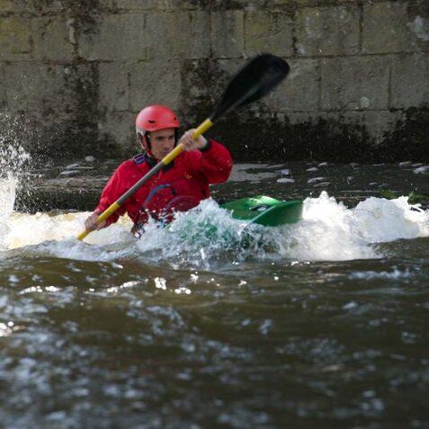 Paddler in roter Ausr&uuml;stung man&ouml;vriert ein Kajak durch spritzendes Wasser an einem Flussufer.