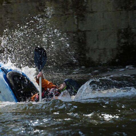 Ein Kajakfahrer kentert mit seinem blauen Kajak in einem sprudelnden Gew&auml;sser. Spritzer fliegen auf.