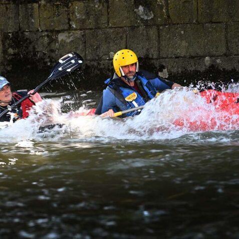 Zwei Personen im Kajak paddeln in einem schnellen Fluss, Spritzer spr&uuml;hen aus dem Wasser.