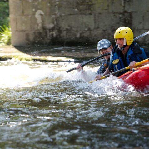 Zwei Kajakfahrer paddeln in einem roten Kajak &uuml;ber eine bewegte Wasserfl&auml;che unter einer Br&uuml;cke.