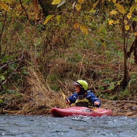 Junger Kajakfahrer in einem roten Boot paddelt auf einem flie&szlig;enden Gew&auml;sser, umgeben von herbstlichem Uferbewuchs.