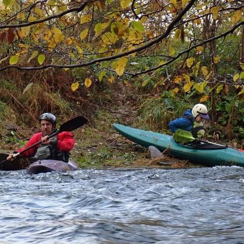 Zwei Kajakfahrer an einem Flussufer, umgeben von herbstlichen Bl&auml;ttern und flie&szlig;endem Wasser.