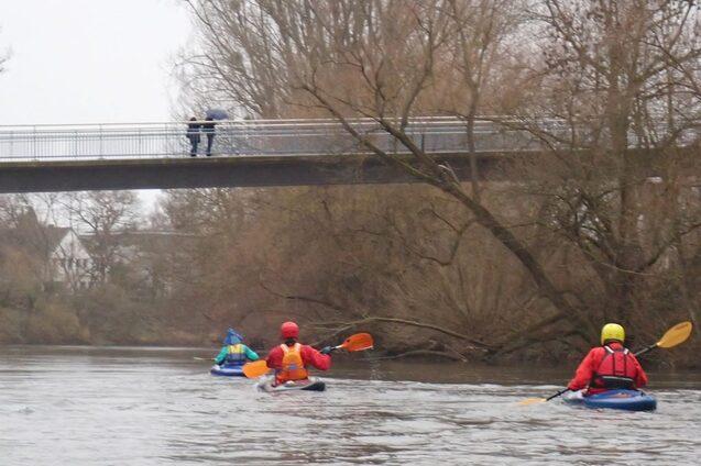 Vier Kajakfahrer paddeln auf einem ruhigen Fluss unter einer Br&uuml;cke, w&auml;hrend zwei Personen darauf stehen.