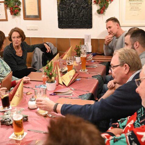 Eine Gruppe von Menschen sitzt an einem Tisch, unterh&auml;lt sich und trinkt Bier in einem festlich dekorierten Raum.