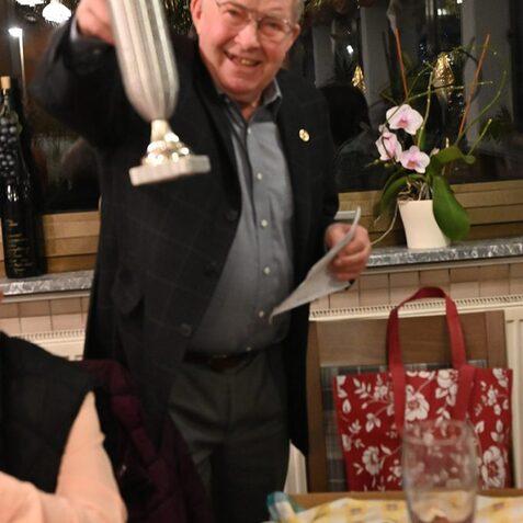 L&auml;chelnder Mann pr&auml;sentiert einen Pokal in einem festlich dekorierten Raum mit Pflanzen und Tischgedeck.