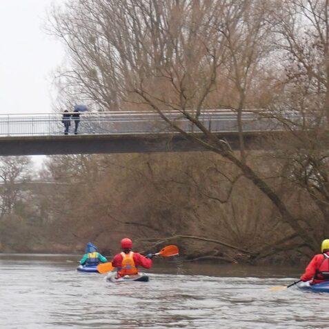 Vier Kajakfahrer paddeln auf einem ruhigen Fluss unter einer Br&uuml;cke, w&auml;hrend zwei Personen darauf stehen.