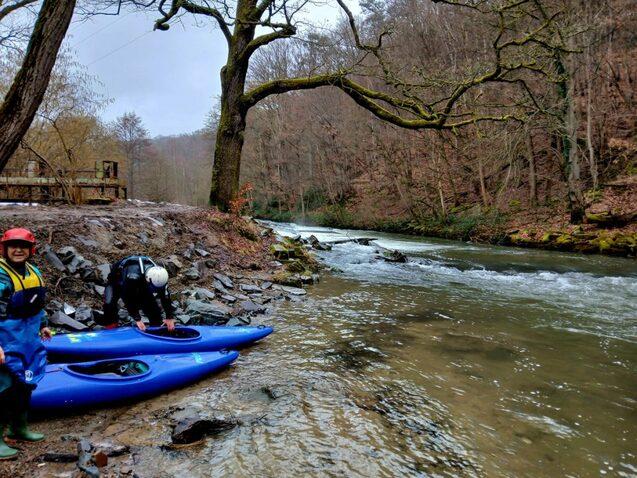 Zwei Personen bereiten ihre Kajaks am Ufer eines flie&szlig;enden Flusses in einem bewaldeten Gebiet vor.
