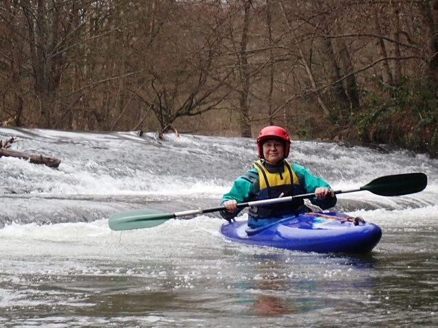 Junge f&auml;hrt mit einem blauen Kajak auf einem ruhigen Fluss, umgeben von B&auml;umen und einer kleinen Wasserfallstelle.