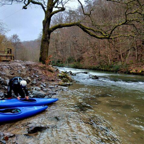 Zwei Personen bereiten ihre Kajaks am Ufer eines flie&szlig;enden Flusses in einem bewaldeten Gebiet vor.