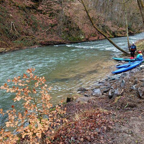 Zwei Kajaks am Ufer eines Flusses, daneben ein Mann mit Ausr&uuml;stung und B&auml;ume entlang der Ufervegetation.