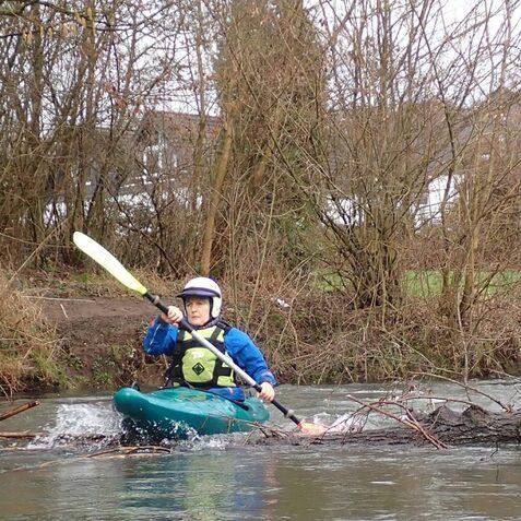 Kajakfahrer paddelt auf einem Fluss und navigiert durch &Auml;ste in der N&auml;he des Ufers.